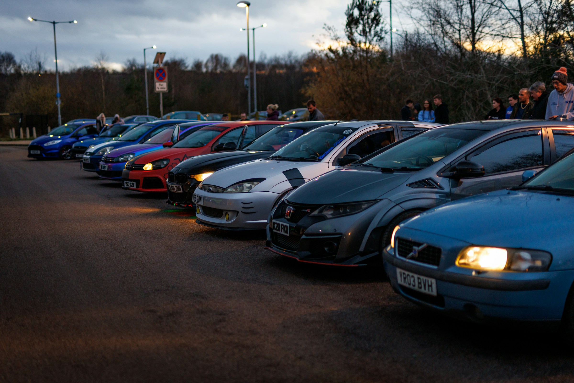 A row of modified hatchbacks lined up at a UK car meet at dusk, with their owners chatting behind them
