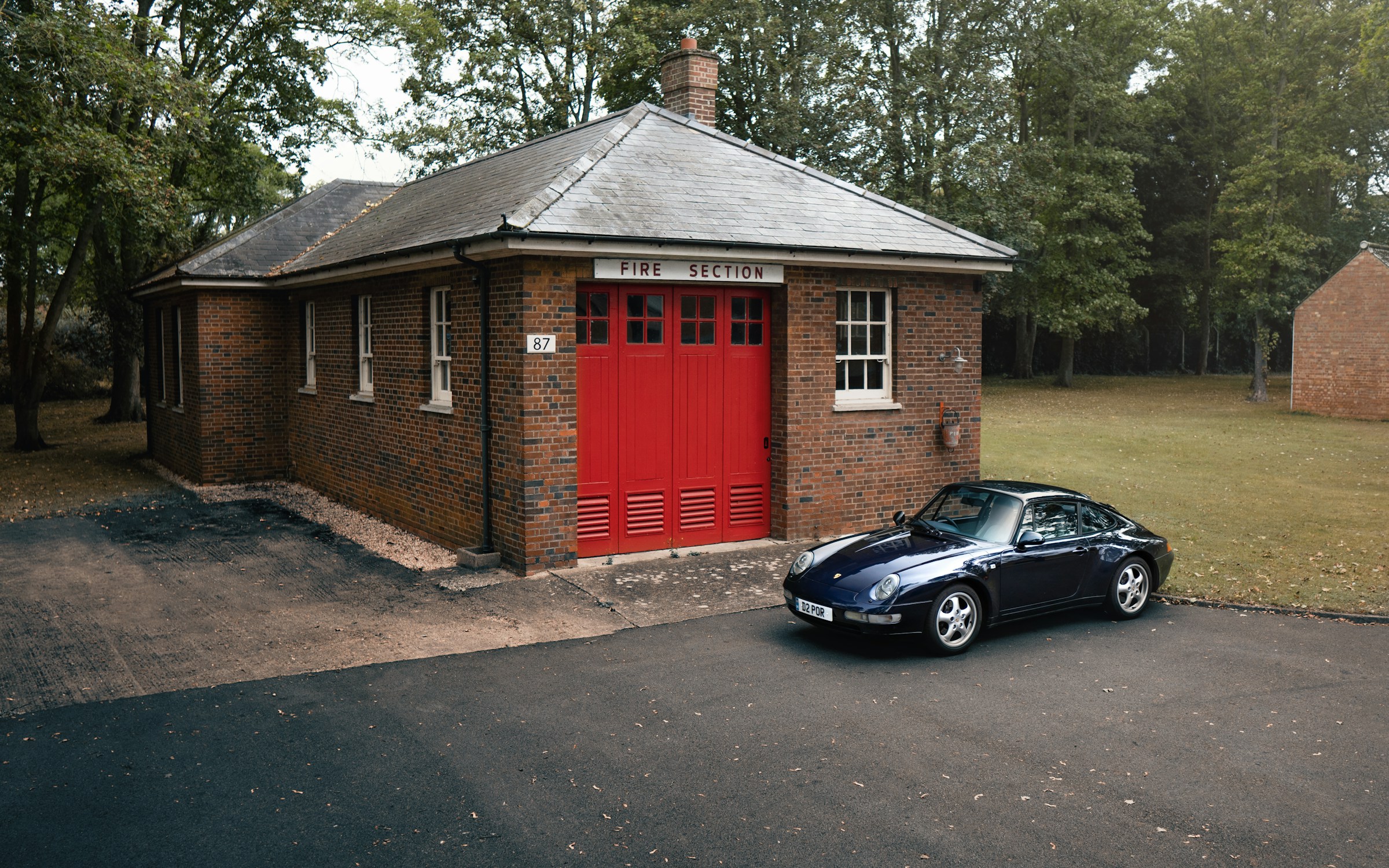 A dark blue Porsche 911 parked outside a red-doored brick fire station at Bicester Heritage, Oxfordshire